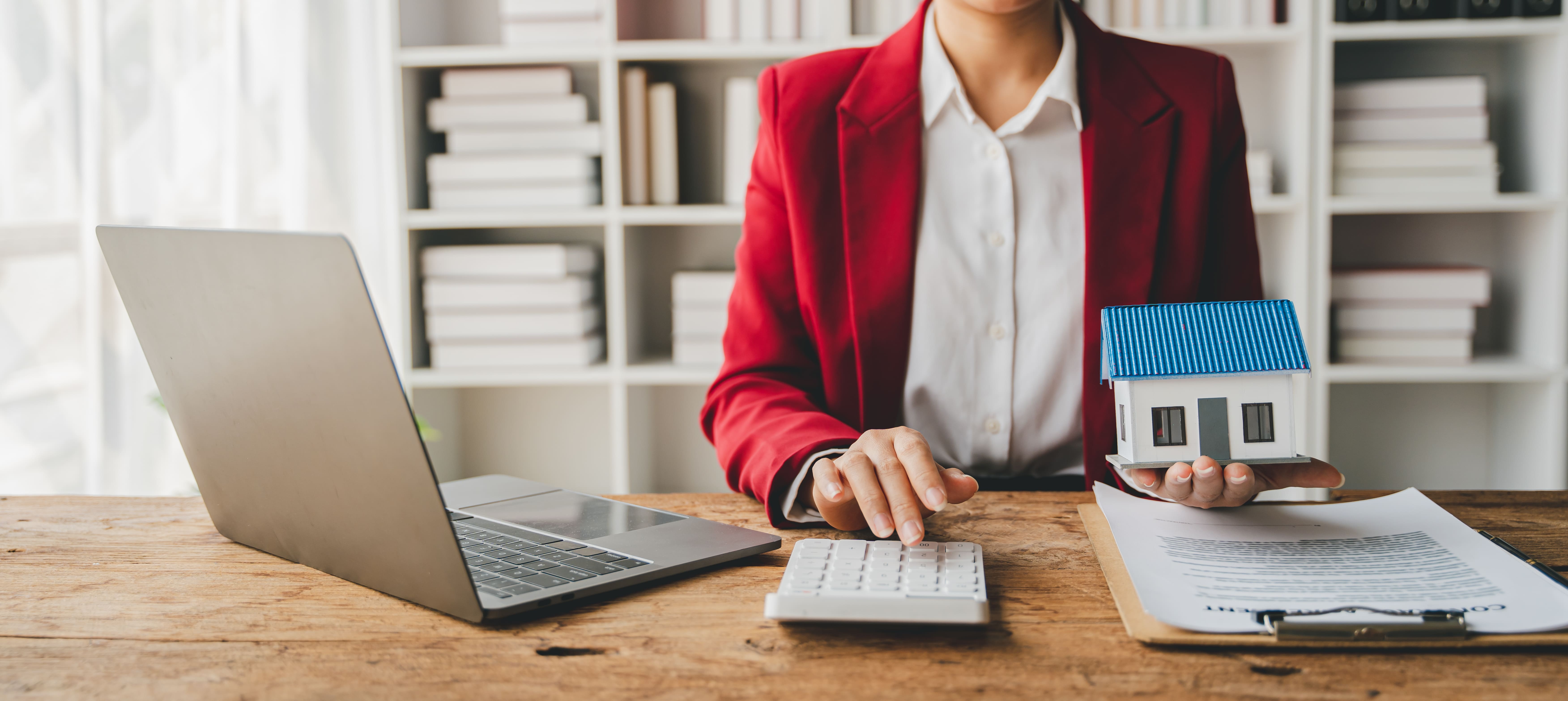 Agent in red blazer holding a house model with a laptop on the desk