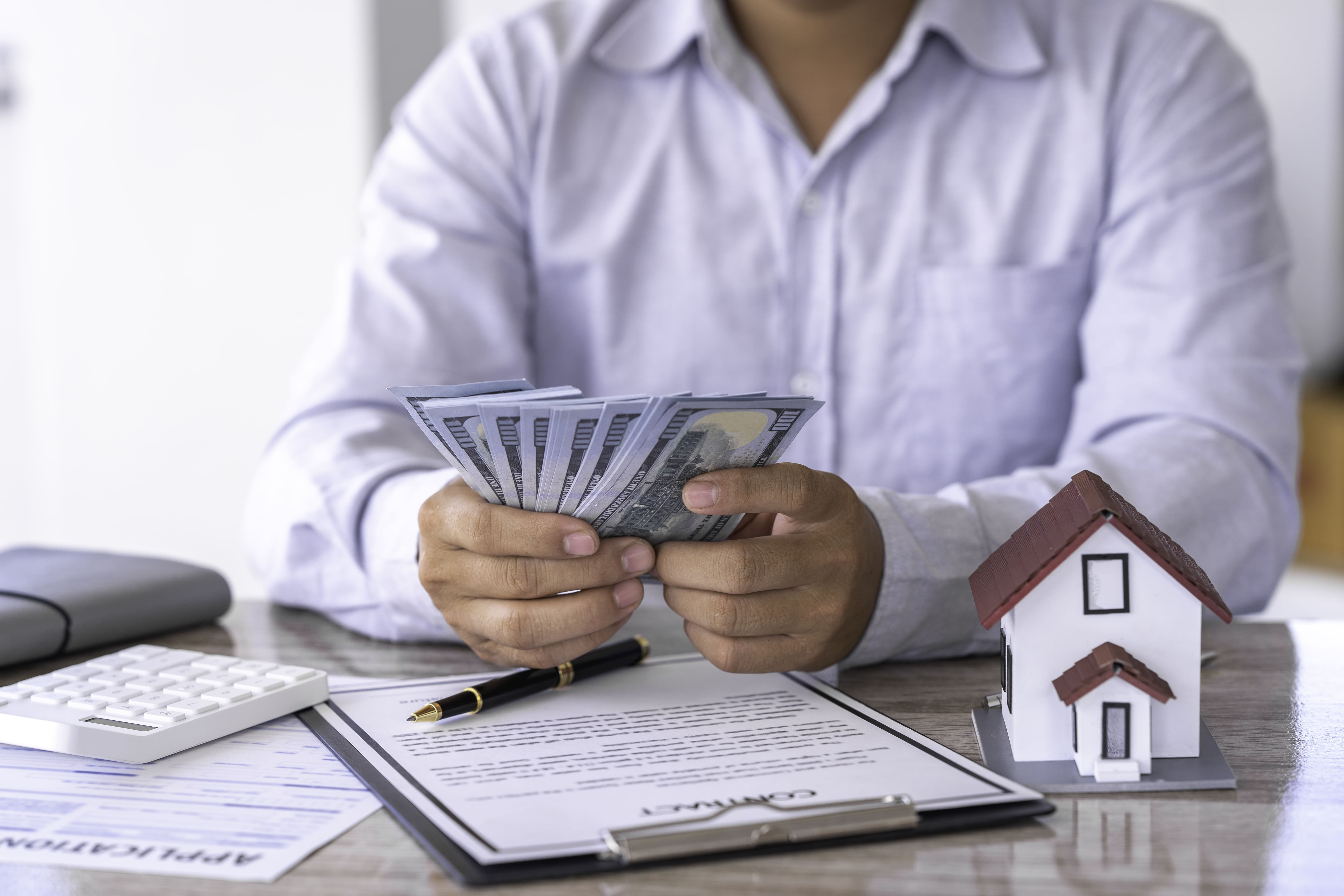 Broker counting cash next to a contract and house model on a desk